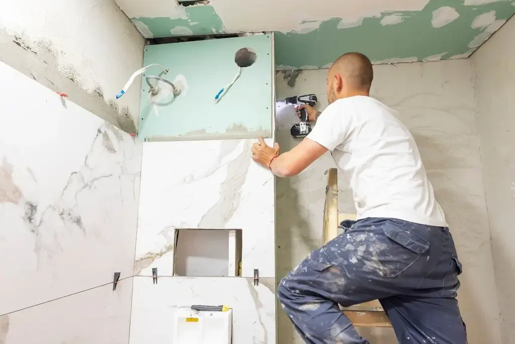 Worker installing large marble-look tiles in a bathroom, standing on a ladder, drilling into wall with exposed plumbing and wiring visible. Renovation in progress.