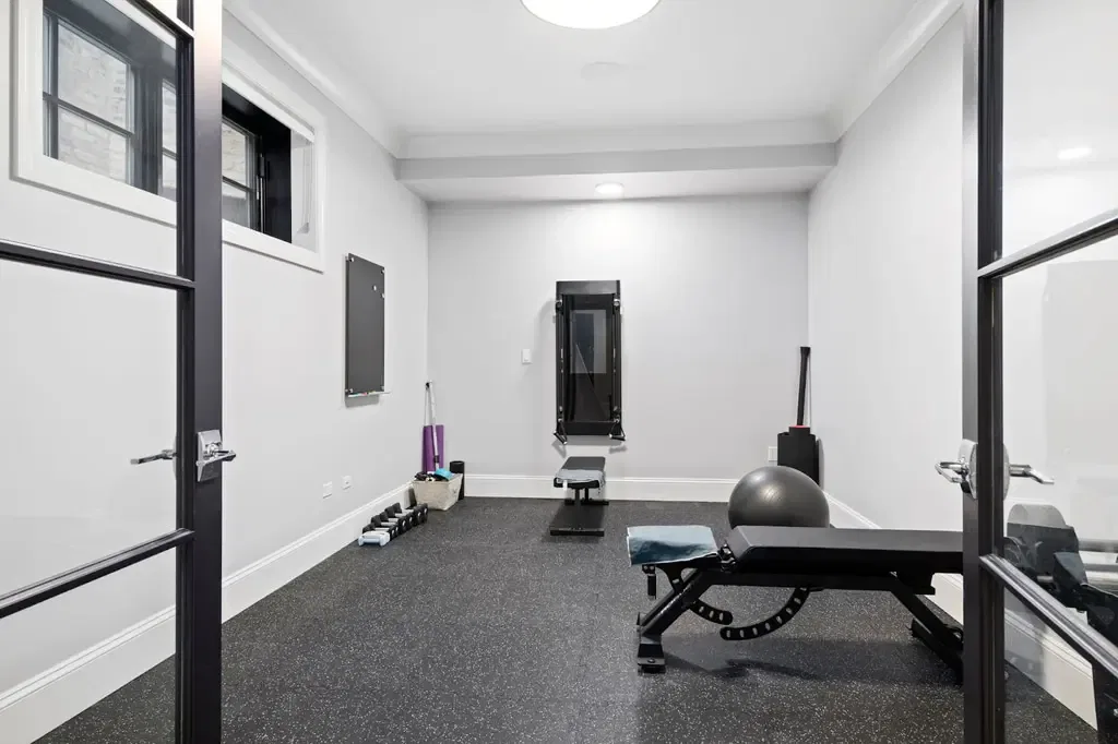 A home gym features black rubber flooring, a weight bench, dumbbells, a stability ball, and a wall-mounted fitness mirror.