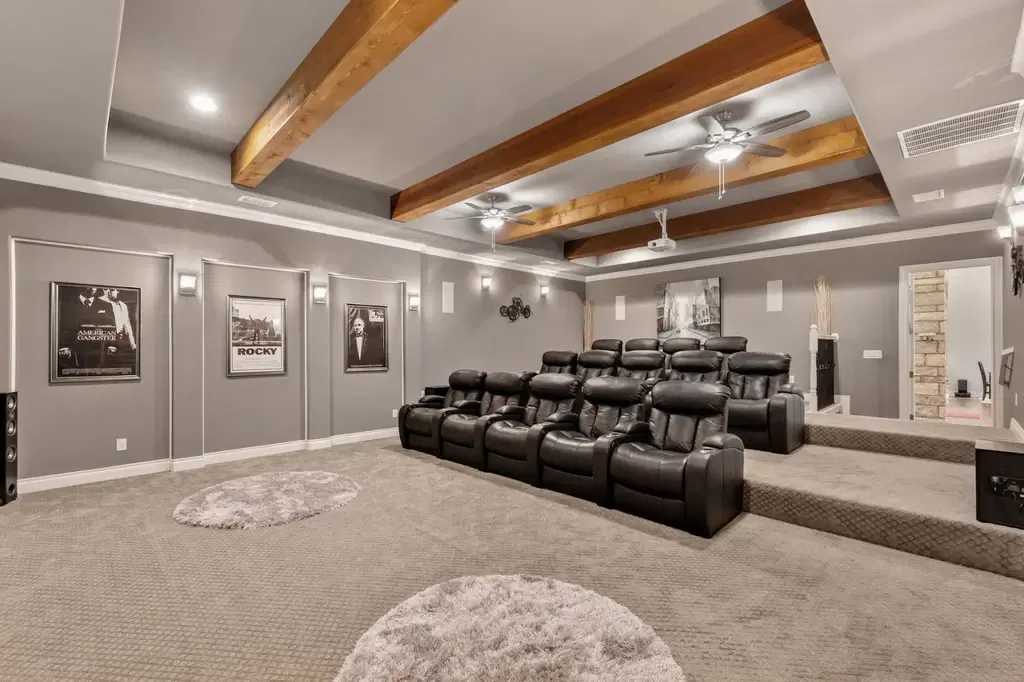 A spacious home theater room features two tiers of black leather reclining armchairs, grey walls adorned with movie posters, and exposed wooden ceiling beams with integrated fans and a projector.